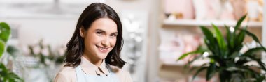 happy florist looking at camera in flower shop on blurred background, banner clipart