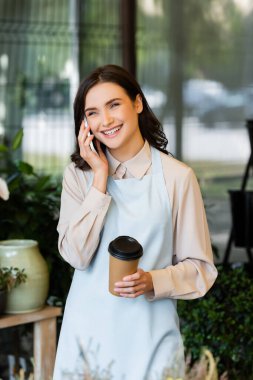 Cep telefonuyla konuşan mutlu çiçekçi ve bulanık ön planda çiçekçide kağıt bardak tutan.