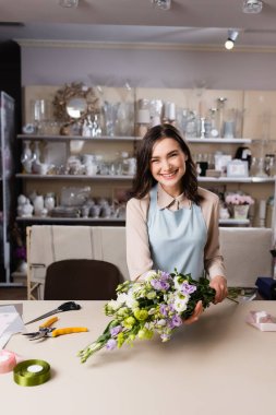 happy florist holding eustoma flowers near rack with vases on blurred background clipart