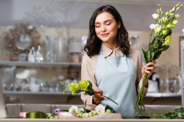 Çiçekçide gülümseyen çiçekçi elinde Eustoma çiçekleriyle ön plandaki bulanık buketi düzenlerken