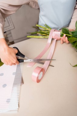 Cropped view of florist with scissors cutting decorative ribbon near tied stalks of bouquet on desk clipart