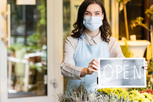 happy florist in medical mask holding board with open lettering near flower shop on blurred background