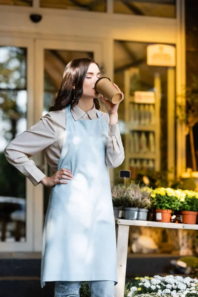 joyful florist holding hand on hip and drinking coffee with closed eyes near flower shop