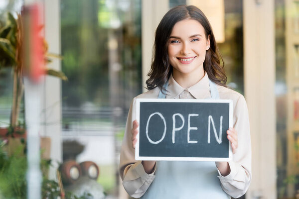 happy florist holding board with open lettering near flower shop while looking at camera