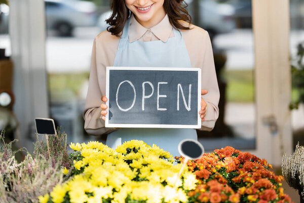 cropped view of smiling florist holding board with open lettering near flowers on blurred foreground