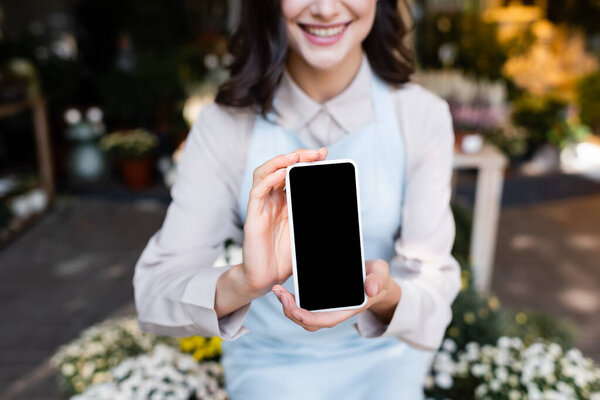 cropped view of smiling florist holding smartphone with blank screen near flowers on blurred foreground