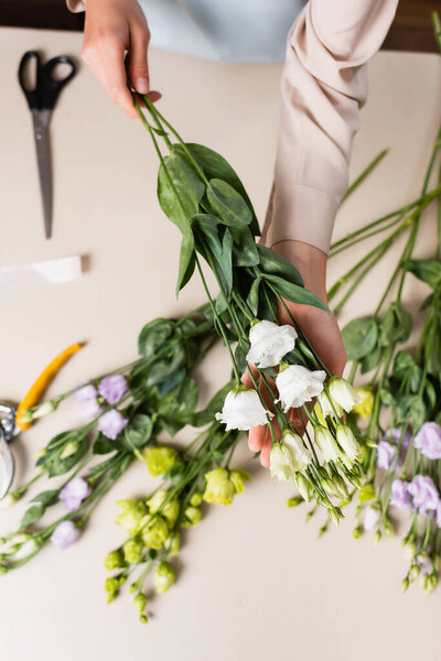 top view of florist holding eustoma flowers near scissors and secateurs while making bouquet on blurred background