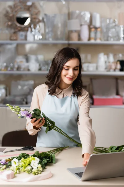 smiling florist holding eustoma flowers while using laptop near rack with vases on blurred background