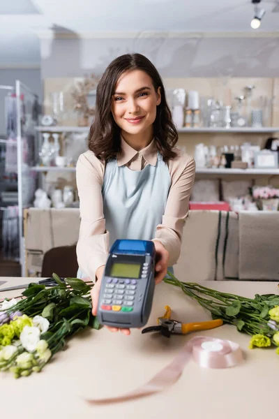 Smiling florist looking at camera while holding terminal near eustoma flowers on table on blurred background