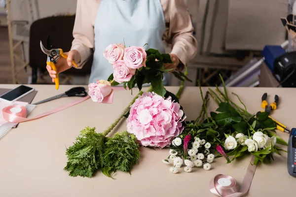 Cropped view of florist holding roses and secateurs near desk with flowers, decorative ribbons and tools on blurred background