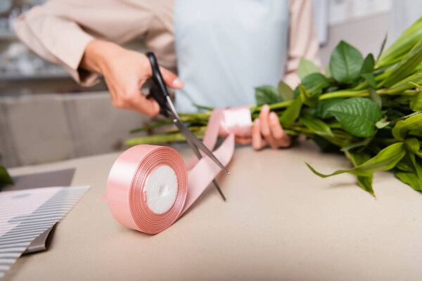 Close up view of decorative ribbon roll near florist with scissors and tied bouquet on desk on blurred background