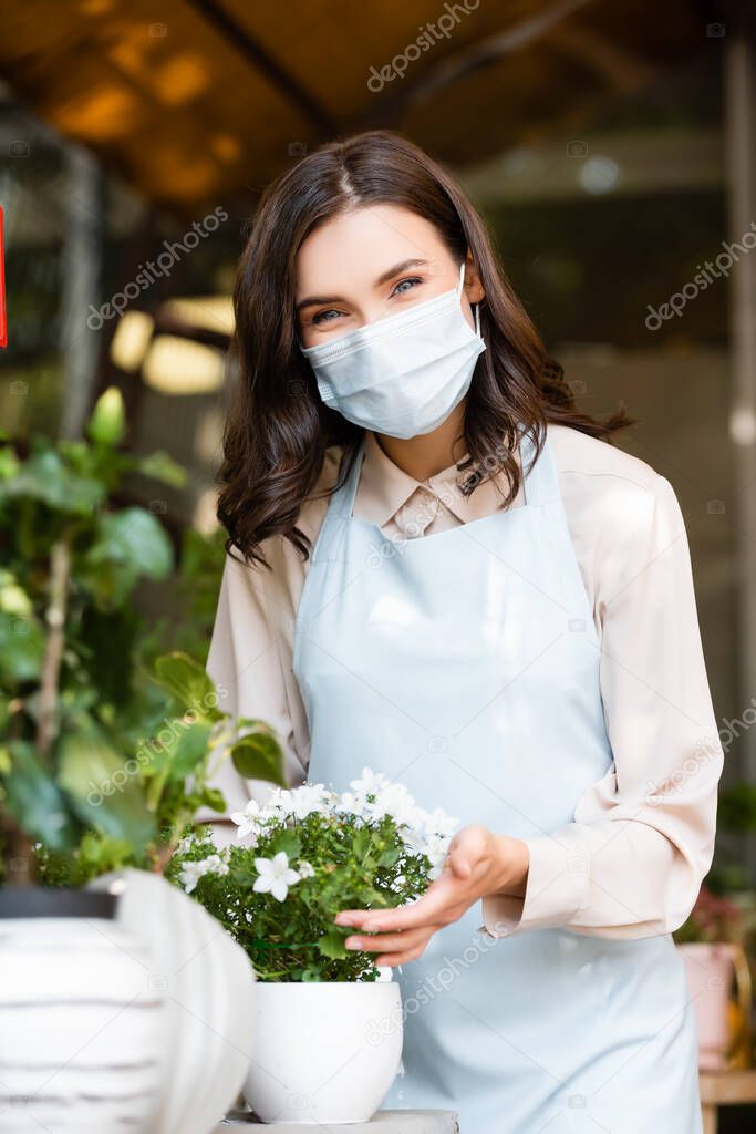 cheerful florist in medical mask caring about plants in flower shop on blurred foreground