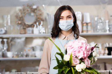 Tıbbi maskeli esmer kadın çiçekçi çiçek dükkanında buketli ve arka planda bulanık askılar var.