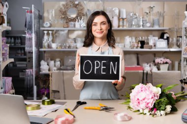 Smiling florist holding chalkboard with open lettering near bouquet on desk with tools and decorative ribbons clipart