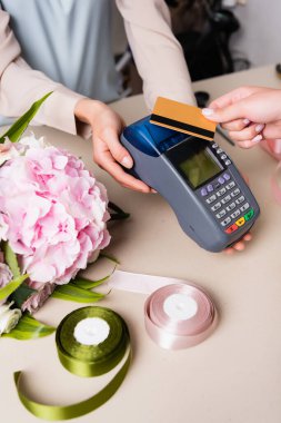Cropped view of customer paying with credit card by terminal in hands of florist near hydrangea and decorative ribbons on desk clipart