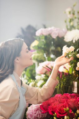 Gülümseyen esmer çiçekçinin yan görünüşü. Bulanık arka plandaki gülleri önemseyen çiçekçi.
