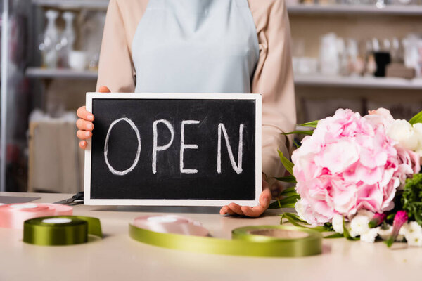 Cropped view of florist holding chalkboard with open lettering near bouquet on desk with blurred decorative ribbons on background