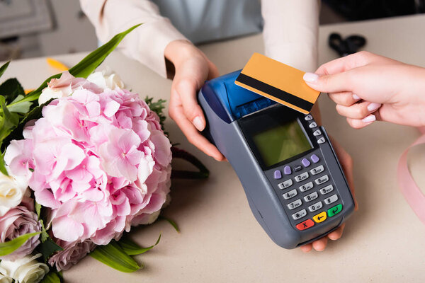 Cropped view of customer paying with credit card by terminal in hands of florist near blooming hydrangea on desk