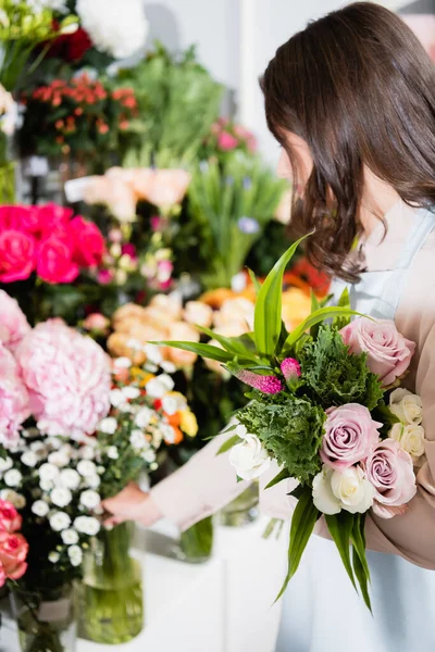 Brunette female florist with bouquet taking branch of chrysanthemums from racks of flowers on blurred background