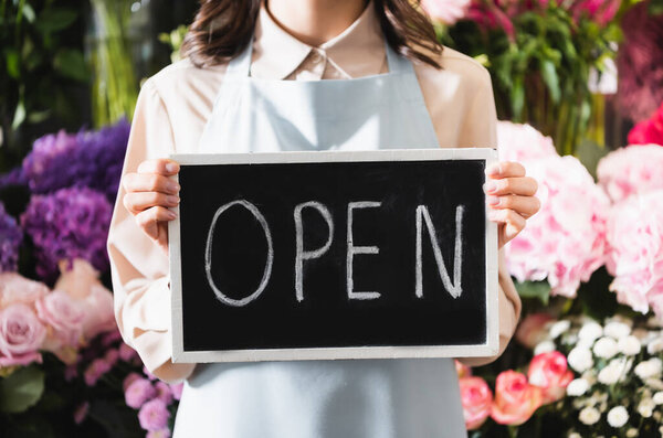 Cropped view of florist holding chalkboard with open lettering with blurred range of flowers on background