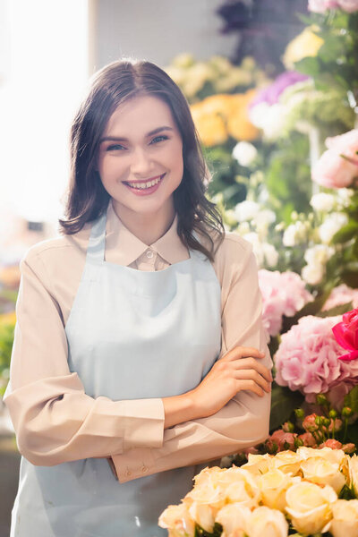 Cheerful female florist with crossed arms looking at camera near racks of flowers on blurred background
