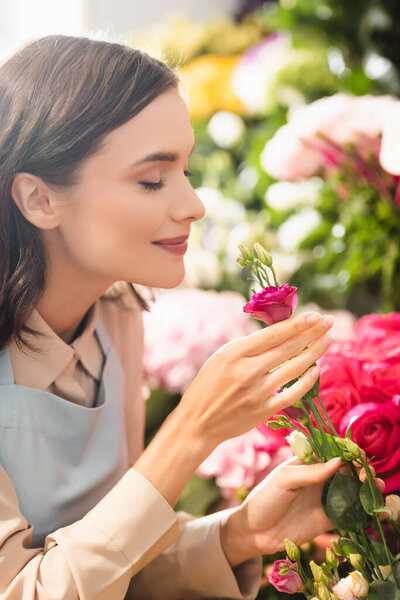 Brunette florist with closed eyes smelling eustoma flower near roses on blurred background