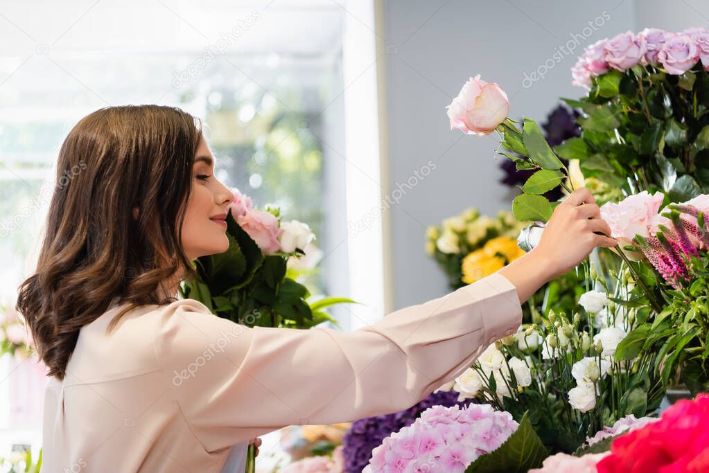 Side view of smiling florist taking rose in store, while arranging bouquet with blurred window on background