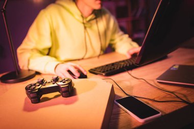 KYIV, UKRAINE - 21 AĞUSTOS 2020: Akıllı telefon ve oyuncunun yanındaki pizza kutusunun üzerindeki joystick bulanık arka planda bilgisayar kullanıyor 