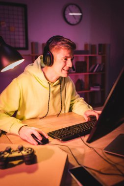 KYIV, UKRAINE - AUGUST 21, 2020: Smiling gamer in headset using computer near smartphone, pizza box and joystick on blurred foreground  clipart