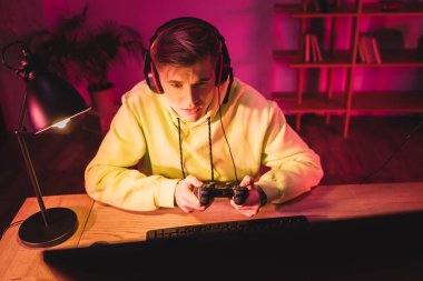 KYIV, UKRAINE - AUGUST 21, 2020: Young gamer in headset using joystick near computer on blurred foreground  clipart