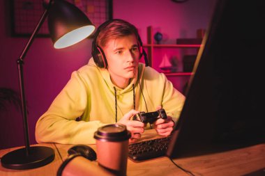 KYIV, UKRAINE - AUGUST 21, 2020: Focused player in headset using joystick near takeaway coffee and computer on blurred foreground  clipart