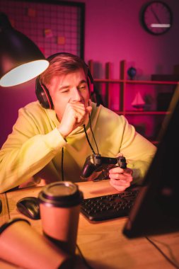 KYIV, UKRAINE - 21 AĞUSTOS 2020: Kulaklık takan yorgun oyuncu bilgisayarın yanında joystick ve kahve kullanarak ön planda bulanıklık yaratıyor 