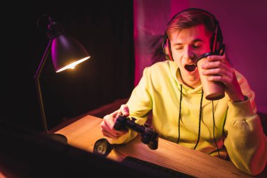 KYIV, UKRAINE - 21 Ağustos 2020: Kulaklık takan oyuncu, ön plandaki bulanık bilgisayarın yanına gitmek için joystick ve kahve tutarken esniyor 