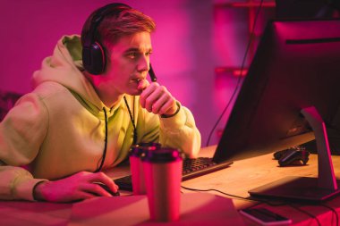 KYIV, UKRAINE - AUGUST 21, 2020: Focused gamer in headset using computer near coffee to go, pizza box and joystick on blurred foreground  clipart