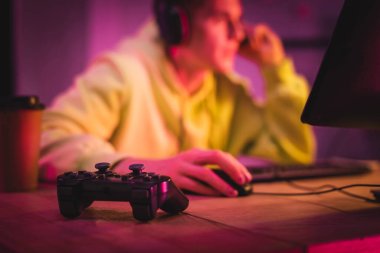 KYIV, UKRAINE - AUGUST 21, 2020: Joystick on table near gamer in headset using computer on blurred background  clipart