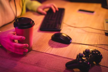 KYIV, UKRAINE - AUGUST 21, 2020: Cropped view of joystick near gamer holding takeaway coffee and using computer keyboard on blurred background  clipart