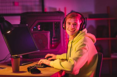 KYIV, UKRAINE - AUGUST 21, 2020: Side view of smiling gamer in headphones using computer keyboard near joystick and coffee to go on blurred foreground  clipart