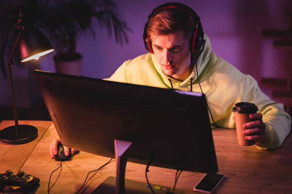 KYIV, UKRAINE - AUGUST 21, 2020: Gamer in headset holding coffee to go near computer, joystick and smartphone on blurred foreground 