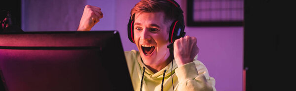 Cheerful gamer in headset showing yeah gesture near computer on blurred foreground, banner 