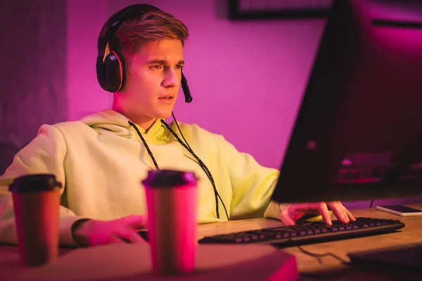 Concentrated player in headset using computer near smartphone and coffee to go on blurred foreground