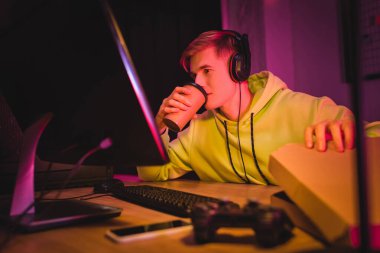 Young gamer in headphones drinking coffee to go and holding pizza box near computer and joystick on blurred foreground  clipart