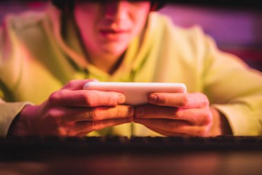 Bulanık arkaplanda bilgisayar klavyesinin yanındaki oyuncunun ellerinde akıllı telefon 