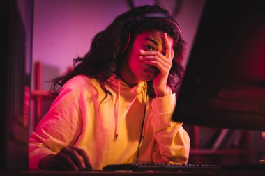 Bulanık ön planda bilgisayar oyunu sırasında bilgisayar kullanarak kulaklık takan yorgun Afrikalı Amerikalı oyuncu 