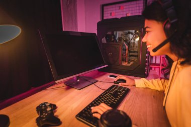 KYIV, UKRAINE - 21 AĞUSTOS 2020: masadaki bulanık ön planda gülümseyen Afro-Amerikan oyuncu, akıllı telefon ve joystick 'in yanında boş ekranlı bilgisayar 