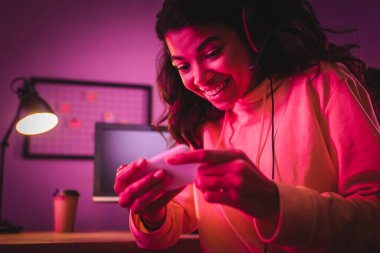 Gülümseyen Afro-Amerikan oyuncu kulaklıkla bulanık ön planda akıllı telefon kullanıyor. 
