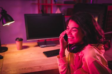 Neşeli Afrikalı Amerikalı oyuncu bilgisayarın yanında akıllı telefondan konuşuyor ve bulanık arka planda kahve içiyor. 