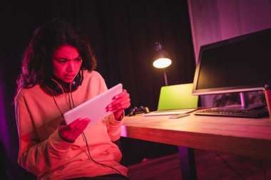 Bulanık arka plandaki cihazların yanında dijital tablet kullanarak kulaklık takan konsantre Afro-Amerikan oyuncu 