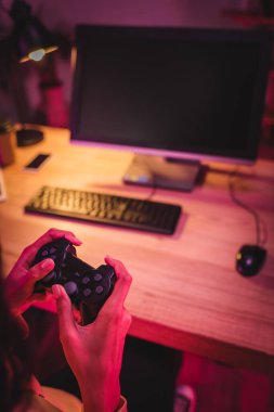 KYIV, UKRAINE - AUGUST 21, 2020: Cropped view of gamer holding joystick near computer on blurred background  clipart