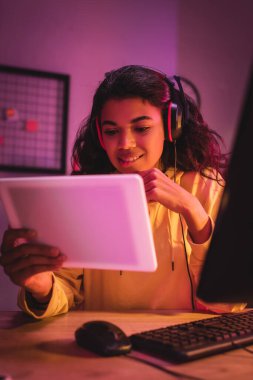 Gülümseyen Afro-Amerikan oyuncu. Kulaklıklı, bulanık ön planda dijital tablet tutuyor. 
