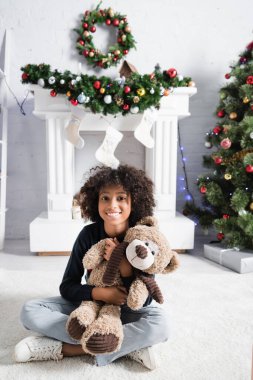 Gülümseyen Afrikalı Amerikalı kız, yerde oyuncak ayıyla otururken kameraya bakıyor.